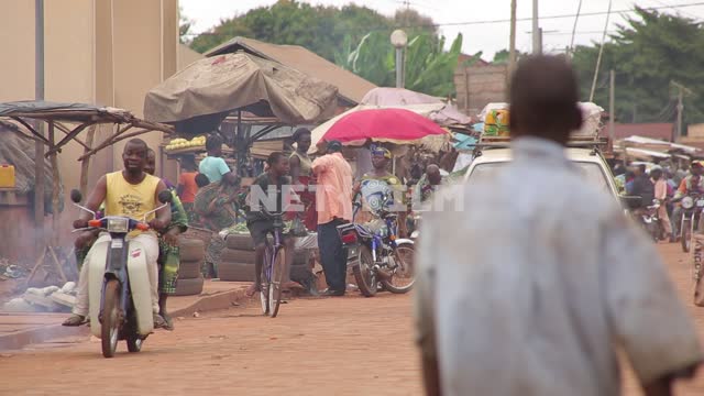Африканский рынок у дороги, африканские люди несут продукты на голове, едут мопеды, мотоциклы,...