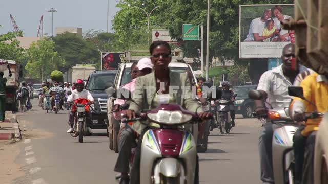 По дороге африканского города едут машины и мотоциклисты, в том числе, в национальных одеждах...
