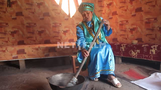 Woman in traditional Altai clothes cook food on the hearth, stirring with a long paddle Woman in...