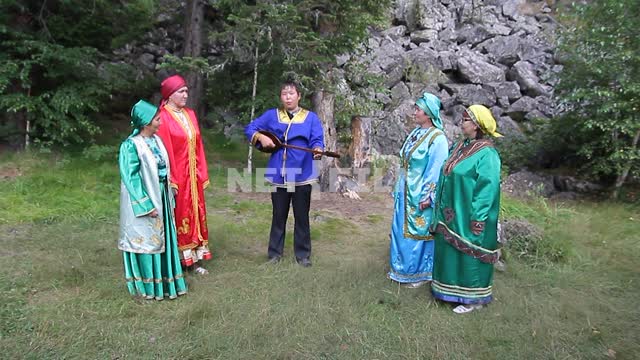 Women in traditional Altai clothes are a circle in a forest clearing around a young musician with...
