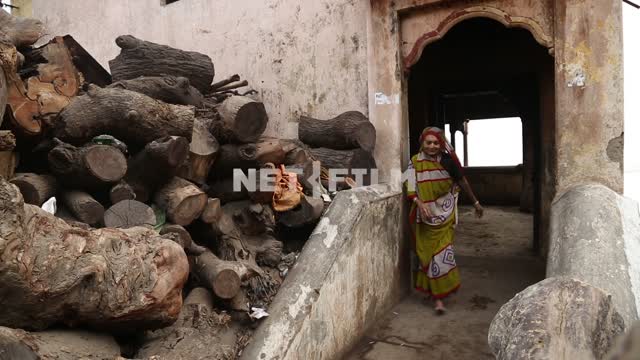 Indian woman comes out of the stone buildings on the street, stops in front of stacked firewood...