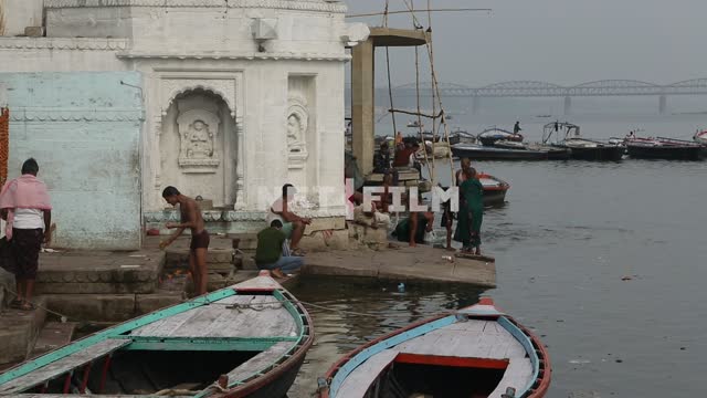 на берегу реки у ступеней индийского храма лодки, индийские мужчины умываются речной водой...