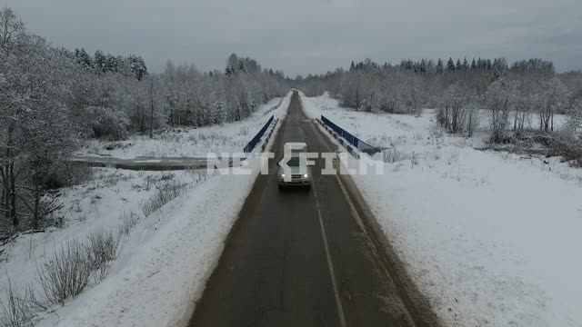 Shooting from above and in front.
Car driving on snowy road, passing the bridge.
The movement of...
