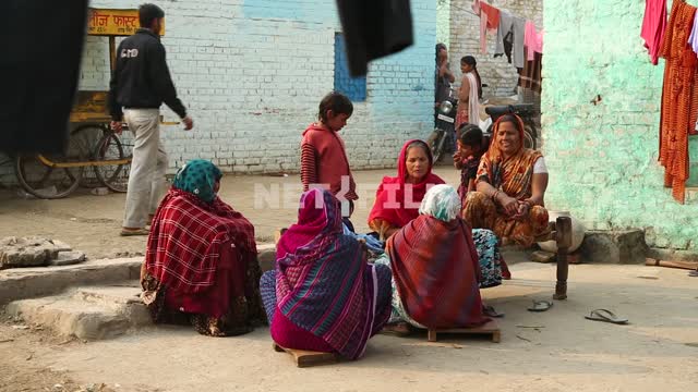 A few Indian women in traditional dress sit outside on a low wooden benches, next hung on a...