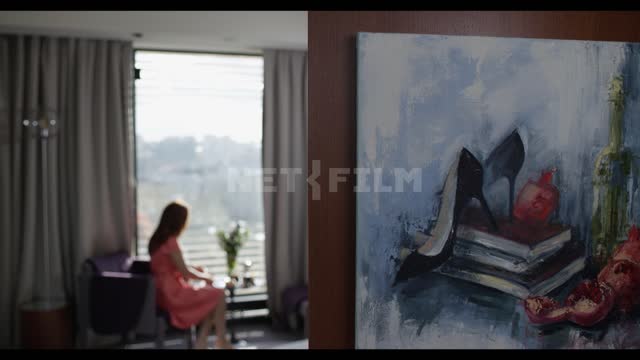 A room with a picture hanging in the foreground, a girl comes to the window, sits in a chair, puts...