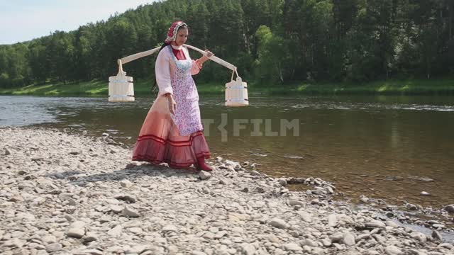 A girl in a national costume walks along the river bank, carries wooden buckets on a yoke Ural,...