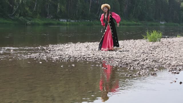 Девушка в национальном костюме танцует на берегу реки, на дальнем плане кто-то бросает камни в воду...
