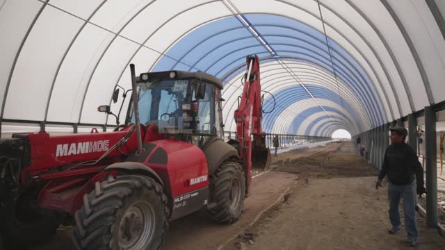 The frame of the hangar is under a blue and white awning, an excavator is working inside, people...