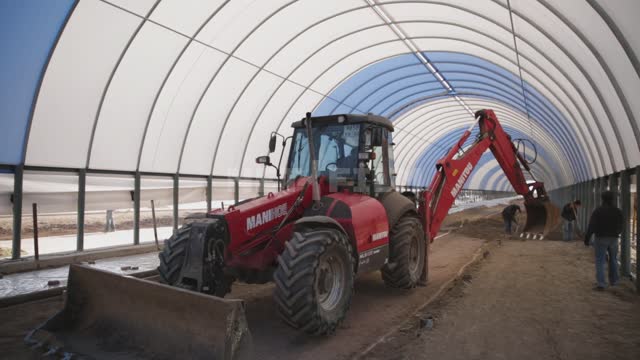 The frame of the hangar is under a blue and white awning, an excavator is working inside Ural,...
