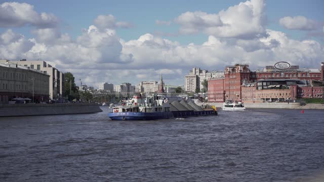 По реке идет грузовая баржа, впереди и навстречу идут прогулочные теплоходы Москва-река, здания,...
