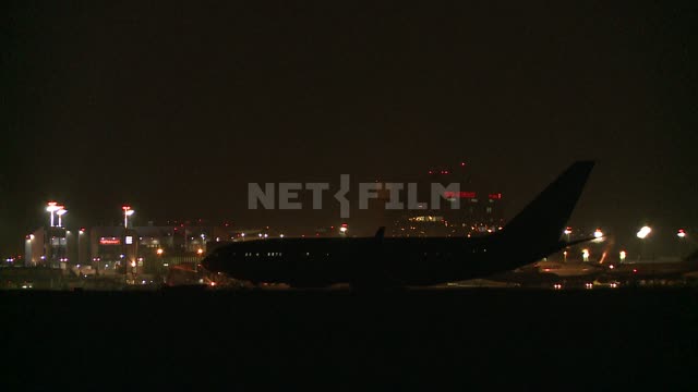 Ночью самолет стоит на взлетной полосе на фоне огней здания аэропорта. Аэропорт, транспорт, взлет,...