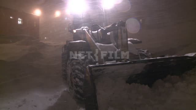 A bulldozer removes snow from the road near the houses by night Russian North, transport, night,...